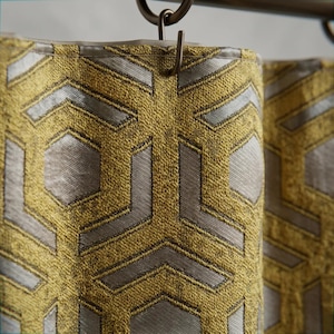 Peut inclure: Un panneau de rideau présentant un motif géométrique répétitif en jaune moutarde et argent métallique. Le tissu possède un tissage texturé et est suspendu à une tringle en métal foncé par une série de crochets métalliques fixés au bord supérieur.