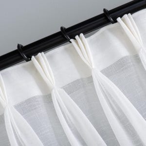 Puede incluir: Primer plano de cortinas de lino blanco con una barra de cortina y anillos negros. Las cortinas están recogidas y atadas a intervalos, creando un aspecto texturizado y elegante. La tela parece semi-transparente.