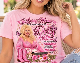 May include: A light pink t-shirt featuring a graphic of Dolly Parton in a pink outfit with a guitar, pink cowboy boots, butterflies, and flowers. The text reads In Loving Memory of a Beautiful Soul Dolly Parton and If you want the rainbow you gotta put up with the rain Forever in our hearts.