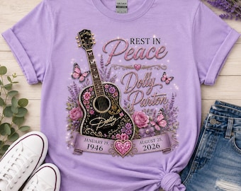May include: A light purple t-shirt featuring a graphic of a black acoustic guitar decorated with pink flowers and butterflies. The text reads "REST IN Peace Dolly Parton" with the dates "JANUARY 19, 1946" and "AUGUST 25, 2026" on a banner.