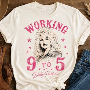 May include: A cream-colored short-sleeve t-shirt featuring a graphic of Dolly Parton with blonde hair. The text reads "WORKING 9 TO 5" in pink distressed lettering, accented by pink stars and the signature "Dolly Parton" with a small heart.