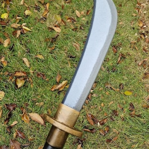 Puede incluir: Una hoja curva de color plateado con una empuñadura dorada y un mango negro. La hoja se encuentra sobre un fondo de hierba verde y hojas caídas, lo que sugiere un entorno al aire libre.