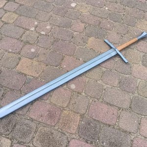 Peut inclure: Une longue épée argentée avec une garde en forme de croix et une poignée enveloppée de couleur beige. L'épée est posée sur une surface en briques.