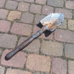 Könnte beinhalten: Eine kleine, antike Schaufel mit einer verwitterten Metallklinge und einem dunklen Holzgriff. Die Schaufel weist Rost- und Abnutzungserscheinungen auf, was darauf hindeutet, dass es sich um ein antikes Stück handelt oder stark genutzt wurde. Das Werkzeug liegt auf einer Ziegeloberfläche.