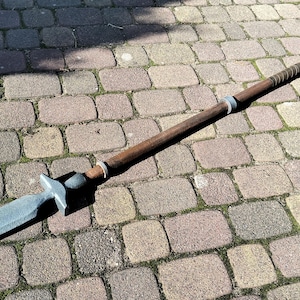 Peut inclure: Une longue lance dotée d'une lame gris métallique pointue fixée à un manche en bois brun foncé. Le manche est orné de plusieurs anneaux argentés et d'une section de prise texturée près de l'extrémité, créant un design équilibré et allongé.