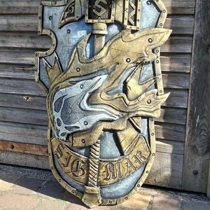 Pode incluir: Um escudo decorativo com um emblema central de martelo com um S estilizado, um motivo de caveira prateada e uma faixa dourada na parte inferior com o texto SIG MAR. O escudo tem um acabamento metálico prateado e dourado com detalhes rebitados.
