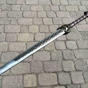 Könnte beinhalten: Dekoratives Schwert mit silberner Klinge und detailliertem Griff. Der Griff ist mit einem dunkelbraunen Material umwickelt. Das Schwert liegt auf einer gepflasterten Oberfläche. Die Klinge hat ein kompliziertes Muster.