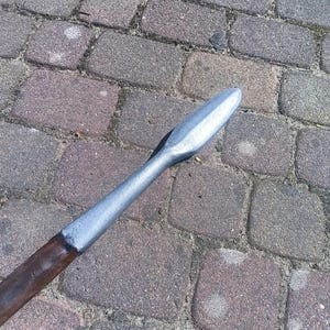 Puede incluir: Una punta de lanza con una hoja plateada y un asta de madera. La punta de lanza es de metal y tiene una punta afilada. El asta es marrón y parece ser de madera. La lanza está sobre una superficie de ladrillo.