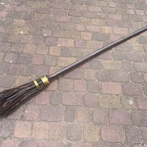 Puede incluir: Una escoba marrón oscuro con un mango largo y recto y un haz de cerdas en el extremo. El mango está acentuado con una banda dorada. La escoba está sobre una superficie de ladrillo.