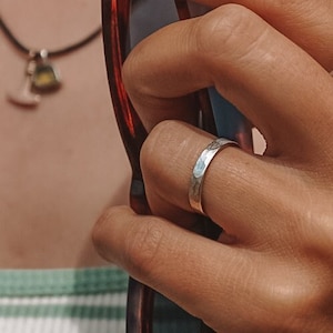 Könnte beinhalten: Ein silberner Ring mit gehämmertem Muster wird an einem Finger getragen. Der Ring ist schlicht und elegant und reflektiert das Licht. Der Hintergrund umfasst eine Halskette und eine Sonnenbrille.