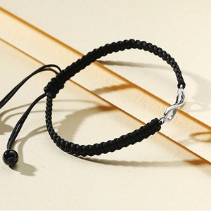 May include: A black braided bracelet with an infinity symbol charm. The bracelet has adjustable ties with black beads at the ends.