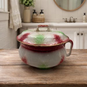 May include: A vintage white porcelain chamber pot with a lid. The pot features a red and green floral design with gold trim. It has a handle on the side and sits on a wooden surface.