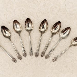 May include: Eight silver spoons arranged in a semi-circle. Each spoon features a long, slender handle with a decorative floral design at the base of the bowl. The spoons are reflective, with a polished finish.