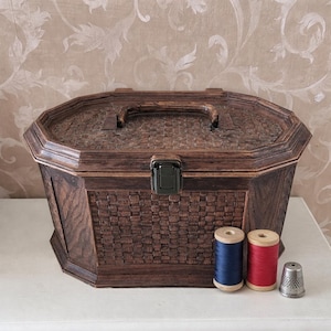Peut inclure: Une boîte à couture en bois brun foncé avec un motif tissé, une poignée et un fermoir noir. Deux bobines de fil, une bleue et une rouge, et un dé à coudre argenté sont devant la boîte. La boîte est octogonale.