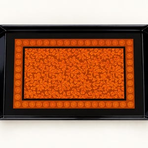 May include: A rectangular black serving tray with integrated side handles. The centre features a vibrant orange panel decorated with a dense, repeating floral leaf pattern, framed by a border of small, circular orange medallions.