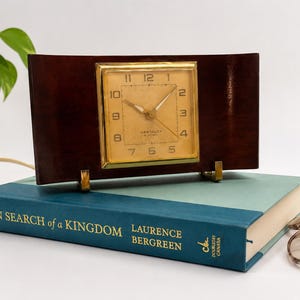 May include: A vintage rectangular clock with a gold-colored face and hands, set in a dark brown wooden frame. The clock is propped up by gold-colored supports and sits atop a teal book. The book's title is "IN SEARCH of a KINGDOM".