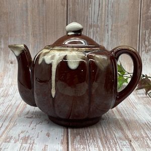 May include: A brown and white ceramic teapot with a drip glaze. The teapot has a rounded body and a spout on the side. The handle is on the opposite side of the spout.