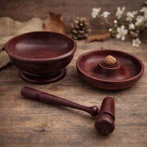 May include: A wooden nutcracker set featuring a bowl, a nut-holding dish, and a mallet. The set is made of dark reddish-brown wood and sits on a wooden surface. A walnut rests in the center of the nut-holding dish. The set is designed for cracking nuts.
