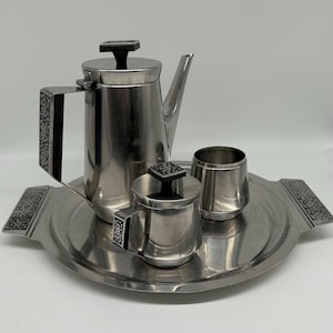 May include: A vintage silver-toned tea or coffee set. The set includes a tall coffee pot, a creamer, a sugar bowl, and a serving tray. The handles and lids have black accents. The tray has decorative detailing on the handles.