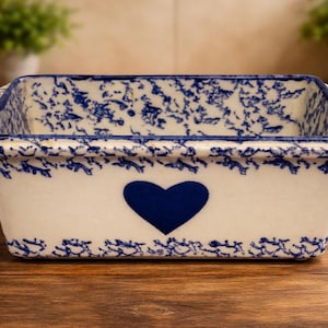 May include: Rectangular ceramic baking dish with a white base and blue floral pattern. A large blue heart is centered on the front. The dish has handles and a blue rim. Ideal for baking and serving.
