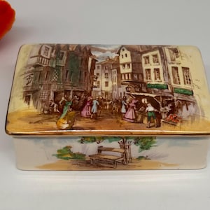 May include: A vintage porcelain trinket box with a colorful scene of a European street with buildings and people. The box has a white base with a floral design and a gold trim.