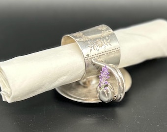 以下が含まれることがあります： ラベンダーの花柄のティーカップの形をした銀製のナプキンリング。リングは白いナプキンを保持しています。