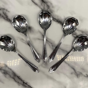 May include: Set of five silver spoons with a shell-shaped bowl. The spoons have a polished finish and are arranged in a fan shape on a marble surface.