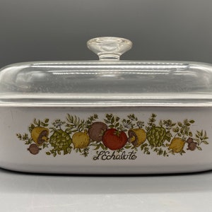 Può includere: Piatto da forno in ceramica bianca con coperchio in vetro. Il piatto presenta un motivo floreale con verdure e la scritta "L'Echalote" stampata sul davanti.