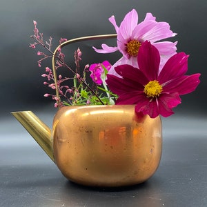 Può includere: Una teiera di rame con un'arrangiamento floreale di cosmos rosa e viola. La teiera ha un manico e un beccuccio dorati.