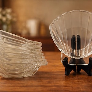 May include: A set of clear glass seashell-shaped serving dishes. One dish is displayed on a black stand, while a stack of similar dishes sits to the left. The dishes are on a wooden surface.