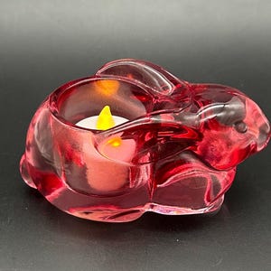 May include: A red glass bunny-shaped candle holder with a battery-operated candle inside. The candle holder is made of transparent red glass and has a smooth, glossy finish. The candle has a yellow flame.