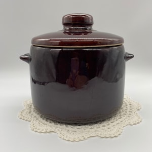 May include: A brown ceramic crock pot with a lid. The crock pot has a rounded shape and two handles on the sides. The crock pot is sitting on a white crocheted doily.