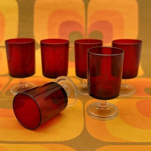 May include: Six vintage red wine glasses with clear stems and bases. The glasses are arranged on a retro orange and yellow patterned surface. One glass is lying on its side, while the others stand upright.