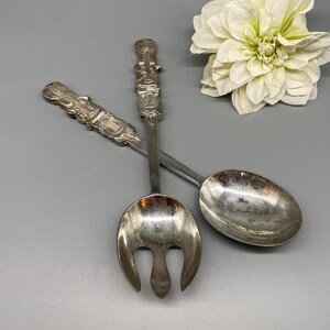 May include: Silver serving utensils with ornate handles, including a fork and spoon. The fork has a three-tine design. The handles feature detailed decorative elements. A white flower is in the background.