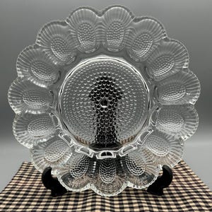 Peut inclure: Plat à servir en verre transparent avec un bord festonné et des empreintes en forme d'œuf. Le centre du plat présente un motif circulaire texturé. Le plat est présenté sur un support noir sur un tissu à carreaux.