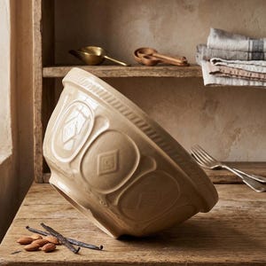 May include: A large, beige ceramic mixing bowl with a glossy finish, featuring a decorative embossed diamond pattern around the exterior and a textured rim. The bowl is tilted on its side, showcasing its rounded shape and sturdy construction.
