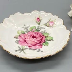 Peut inclure: Un plat à bijoux en porcelaine blanche avec un bord festonné et une bordure dorée. Le plat présente une rose rose avec des feuilles vertes sur un fond blanc.
