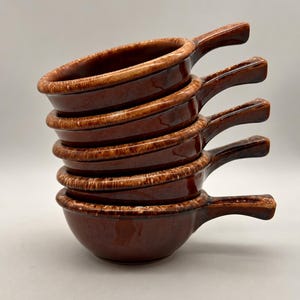 May include: A stack of five round ceramic bowls with handles, each featuring a glossy dark brown glaze with lighter brown mottled accents along the rims and handles. The bowls are arranged vertically, showing a consistent design and finish.