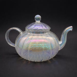 Puede incluir: Una tetera de vidrio transparente con tapa y asa. La tetera tiene un cuerpo redondo con estrías verticales y un brillo nacarado, que refleja los colores del arcoíris. El pico y el asa son curvos.