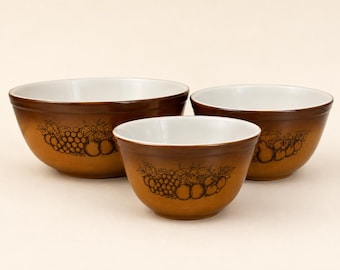 May include: A set of three nesting mixing bowls with a white interior and a brown exterior. Each bowl features a dark brown line art illustration of grapes and other fruits wrapped around the outer surface, creating a consistent visual theme.
