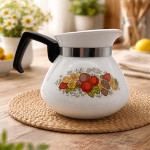 May include: White ceramic teapot with a black handle and a silver band. The teapot features a floral design with yellow, red, and brown elements. It sits on a round, woven placemat on a wooden table.
