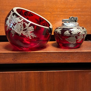 May include: A vintage red glass ashtray and lighter set. The ashtray is a rounded shape, and the lighter is a small, round design. Both pieces feature a silver floral design. The set is displayed on a wooden surface.