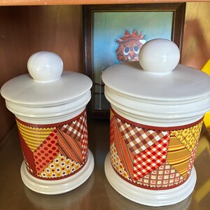 May include: Two cylindrical ceramic canisters with white lids and spherical knobs. The bodies feature a colorful patchwork pattern of orange, yellow, brown, and red fabric-like designs with black stitched details around the edges of each patch.