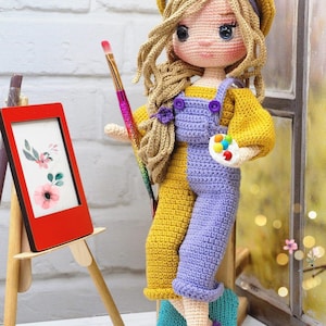 Może przedstawiać: Lalka szydełkowa w żółto-fioletowym stroju, beretce, trzymająca pędzel i paletę. Lalka stoi obok małego sztalugi z obrazem w ramie.