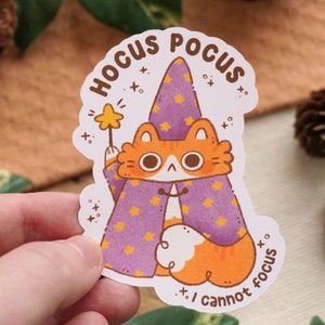 May include: A white sticker with an orange and white cat wearing a purple and yellow wizard hat and holding a wand. The sticker says "Hocus Pocus I cannot focus" in a black font.