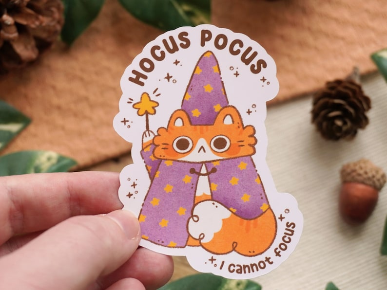 May include: A white sticker with an orange and white cat wearing a purple and yellow wizard hat and holding a wand. The sticker says "Hocus Pocus I cannot focus" in a black font.