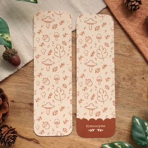 May include: Two brown bookmarks with a white background and a pattern of mushrooms, acorns, leaves, and other autumn-themed elements. The bookmark on the right has the text "bymossypine" at the bottom.