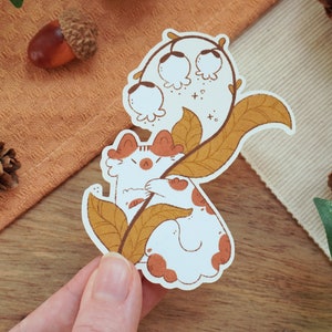 May include: A white cat with orange ears and a brown nose is holding onto a branch of a lily of the valley plant with yellow leaves. The cat is looking to the left. The background is a light brown surface.