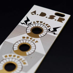 Puede incluir: Panel rectangular blanco con gr&aacute;ficos dorados y negros. El panel presenta el texto "A.D.S.R." en la parte superior, con dise&ntilde;os circulares y las palabras "ATTACK", "DECAY" y "SUSTAIN". Tambi&eacute;n hay gr&aacute;ficos de rayos.