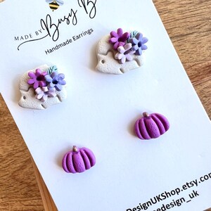 May include: A set of three handmade polymer clay earrings. The earrings feature a white flower design with purple and pink flowers, and two purple pumpkin designs.